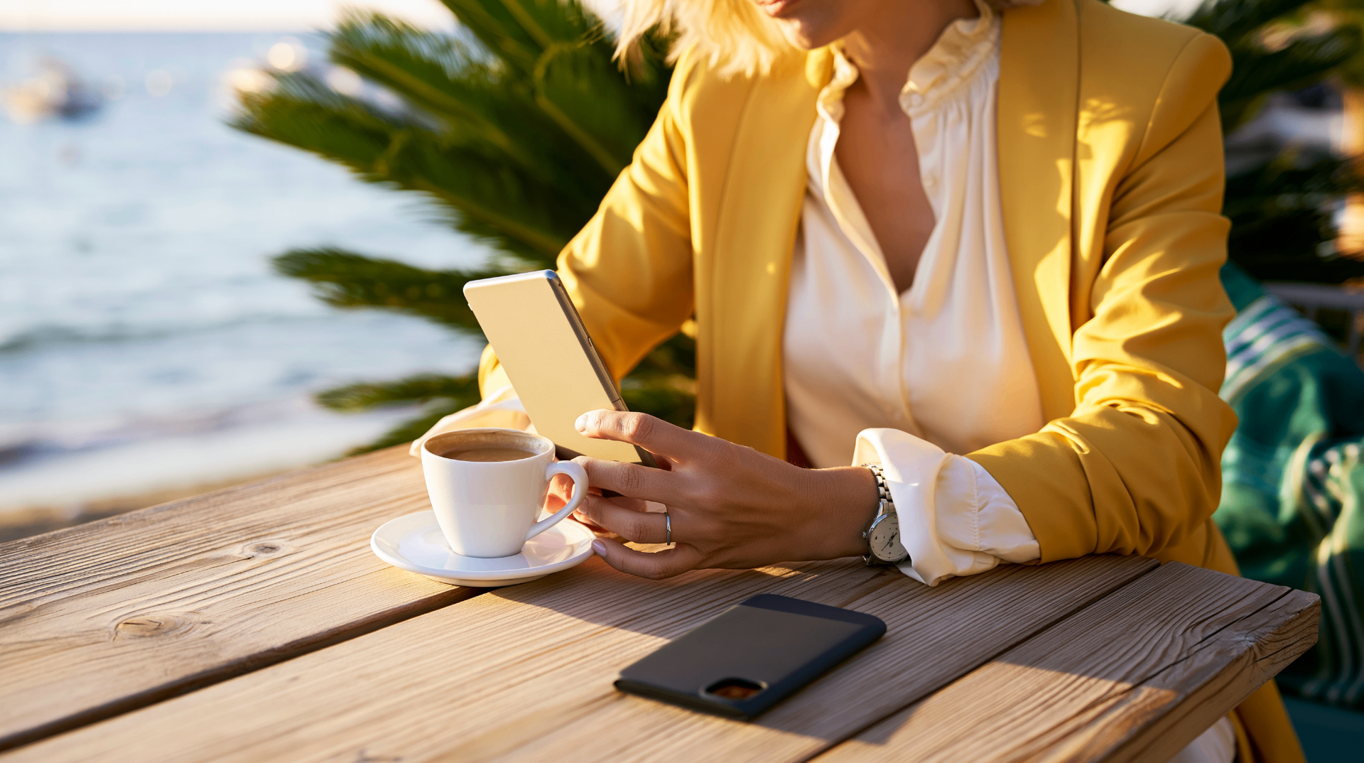Woman in a yellow blazer checking her phone over coffee by the sea