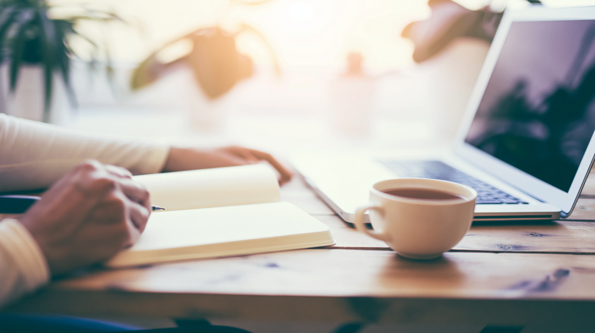 Hands writing in a notebook beside a laptop and coffee